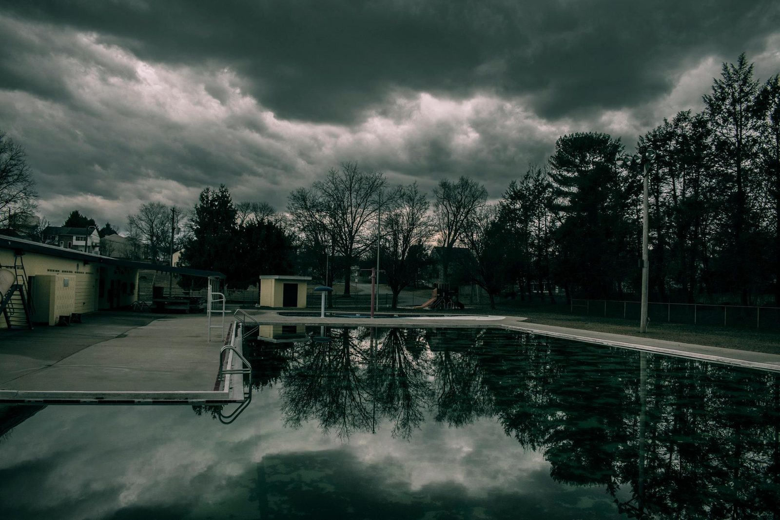 Closed for the Season — Lancaster County. Community pool, no swimmers, storm stacked over the water. The diving board is a line. The sky is doing the diving.